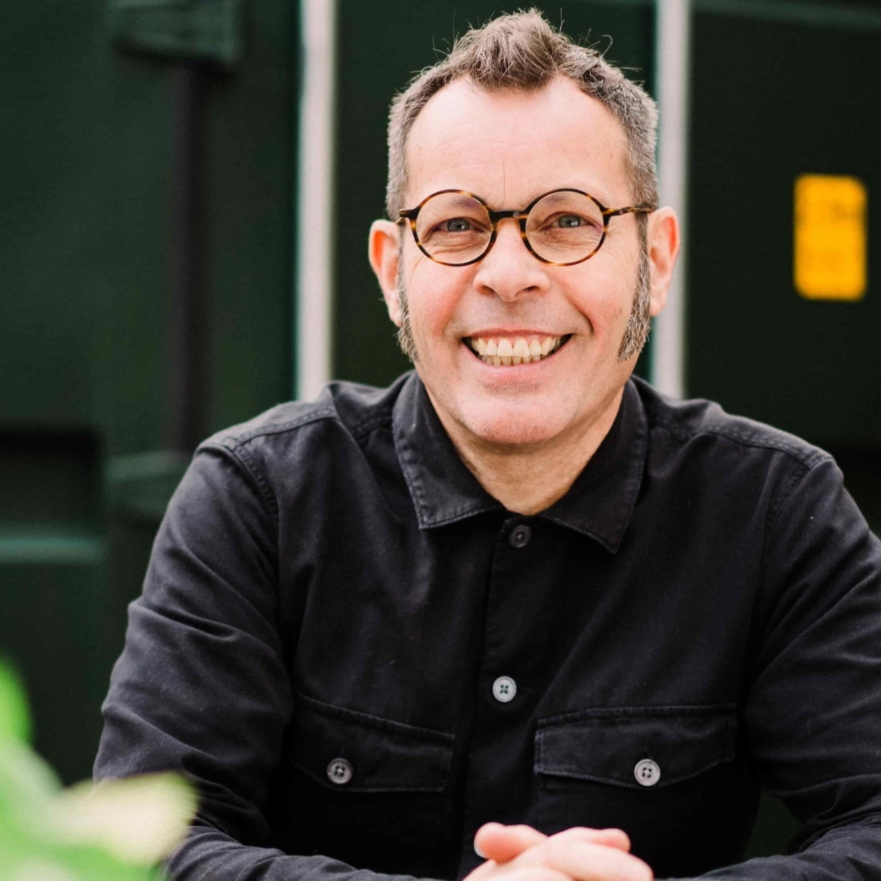 A portrait of a smiling Paul Mackman, a white man with short brown hair and glasses, wearing circular glasses and dark shirt whilst sat at an outdoor table. Behind him is a green shipping metal panel and brickwork